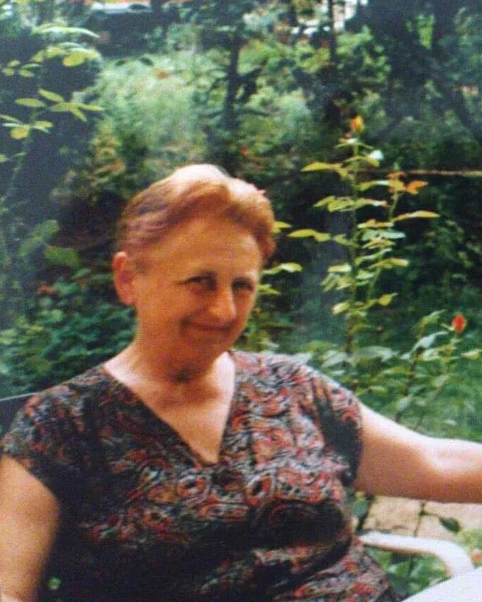 Zorka, an older woman with short reddish hair and a patterned dress, smiling as she sits in a green garden.